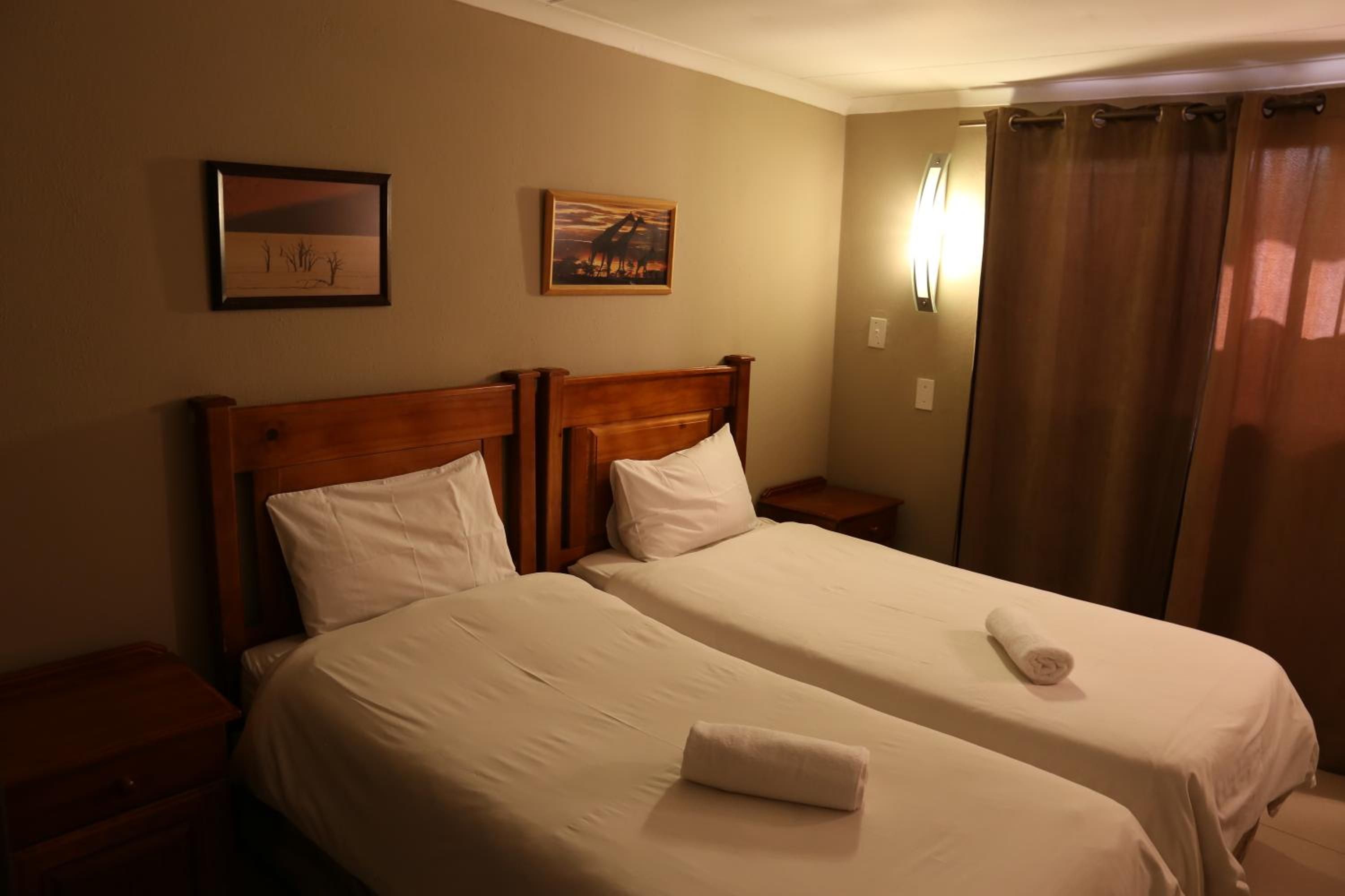 Une chambre d’hôtel avec deux lits, une table de chevet, une tête de lit en bois, une lampe fixée au mur et une fenêtre avec des rideaux.
