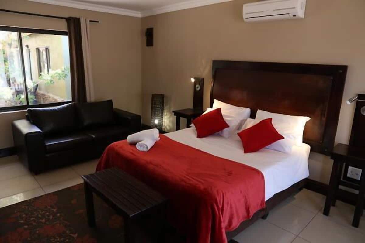 Habitación de hotel con cama, colcha roja, sofá de cuero negro, mesita y ventana con cortinas.
