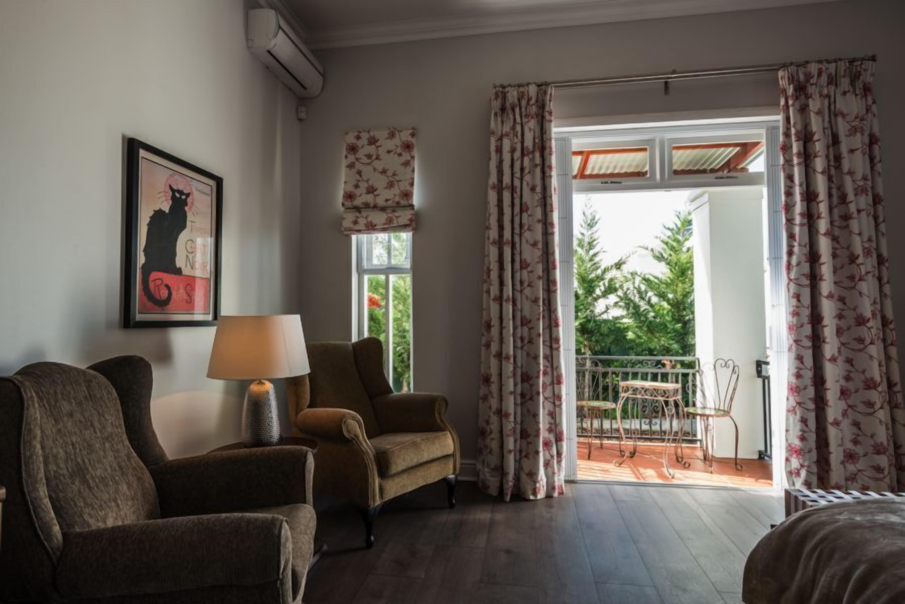 A room with a sofa, armchair, and a table with a lamp. There is a window with curtains and a view of greenery outside.