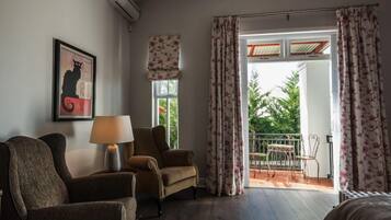 A room with a sofa, armchair, and a table with a lamp. There is a window with curtains and a view of greenery outside.