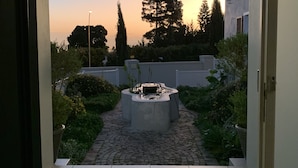 Un arc en pierre mène à un jardin agrémenté d’un foyer, entouré de verdure et surplombé d’un ciel dégagé.