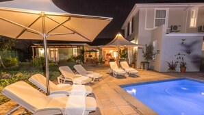 Un espace aménagé au bord de la piscine, avec des chaises longues et des parasols, une piscine, et une maison en arrière-plan.