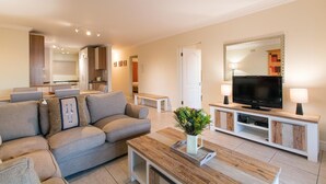 A modern living room with a grey sofa, a wooden coffee table, a TV on a white cabinet, and a kitchen in the background. - Gold Water (Cape Town)
