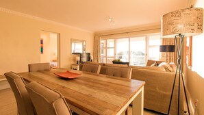 A well-lit living room with a wooden dining table, beige sofas, and a large window providing natural light. - Gold Water (Cape Town)