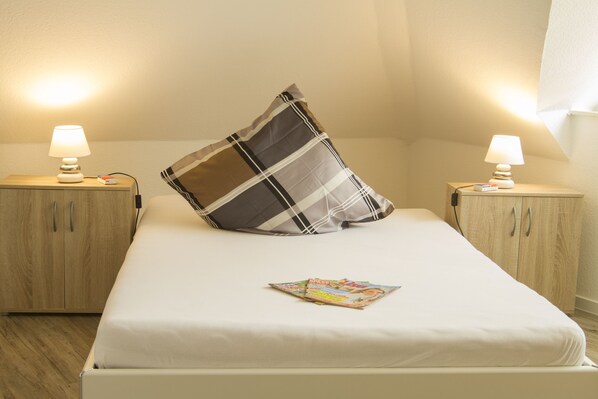 A neatly made bed with a plaid pillow and a magazine on it, flanked by two bedside tables with lamps and a wooden wardrobe. - Haus Betty (Rhauderfehn)