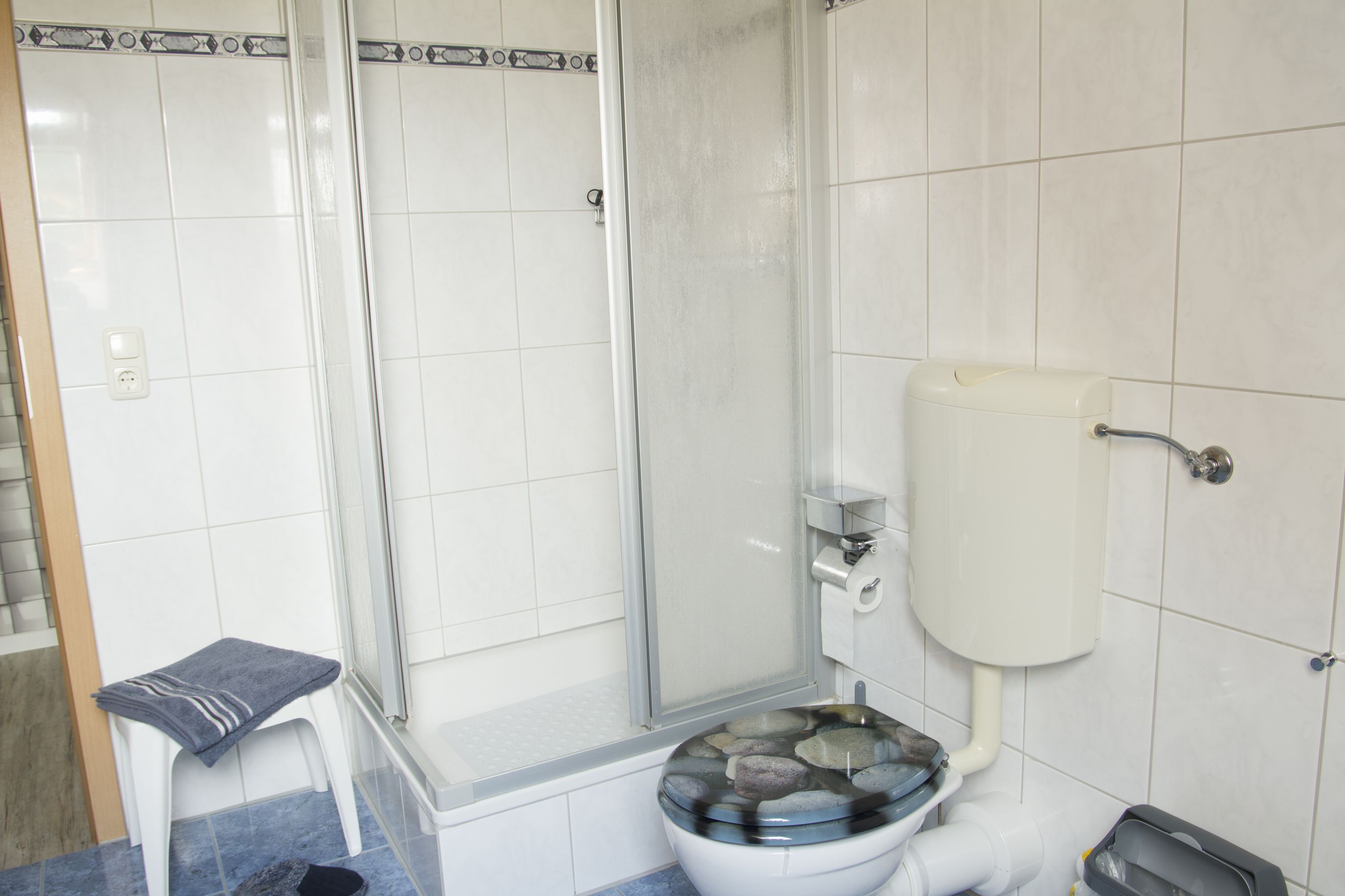Et badeværelse med toilet, brusebad og en lille skammel med et håndklæde.