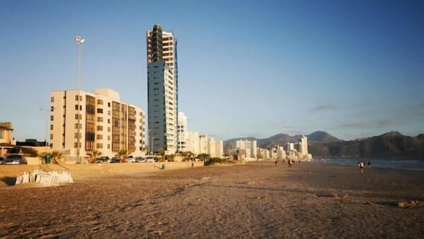 A beach with buildings, including a tall hotel and a shorter building with a sloped roof. - Topaz (Cape Town)