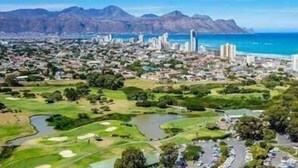 A coastal cityscape with a golf course, residential areas, and a mountain range in the background. - Topaz (Cape Town)