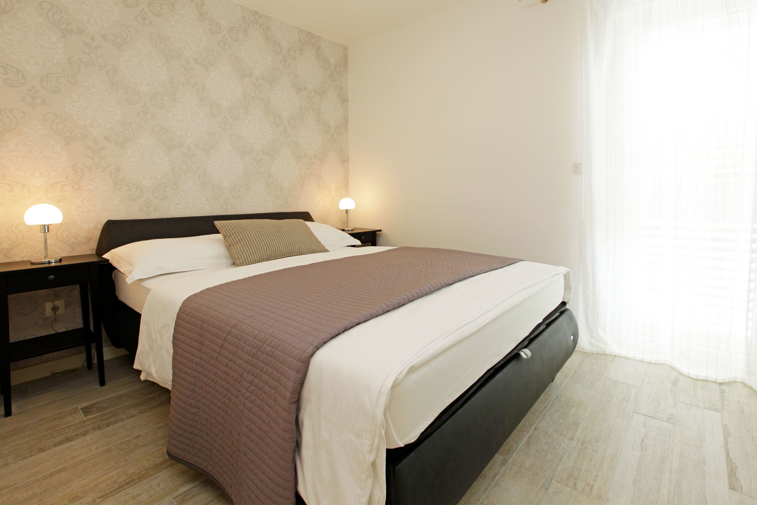 A neatly made bed with a brown and white duvet, two bedside lamps, a nightstand, and a window with sheer curtains.