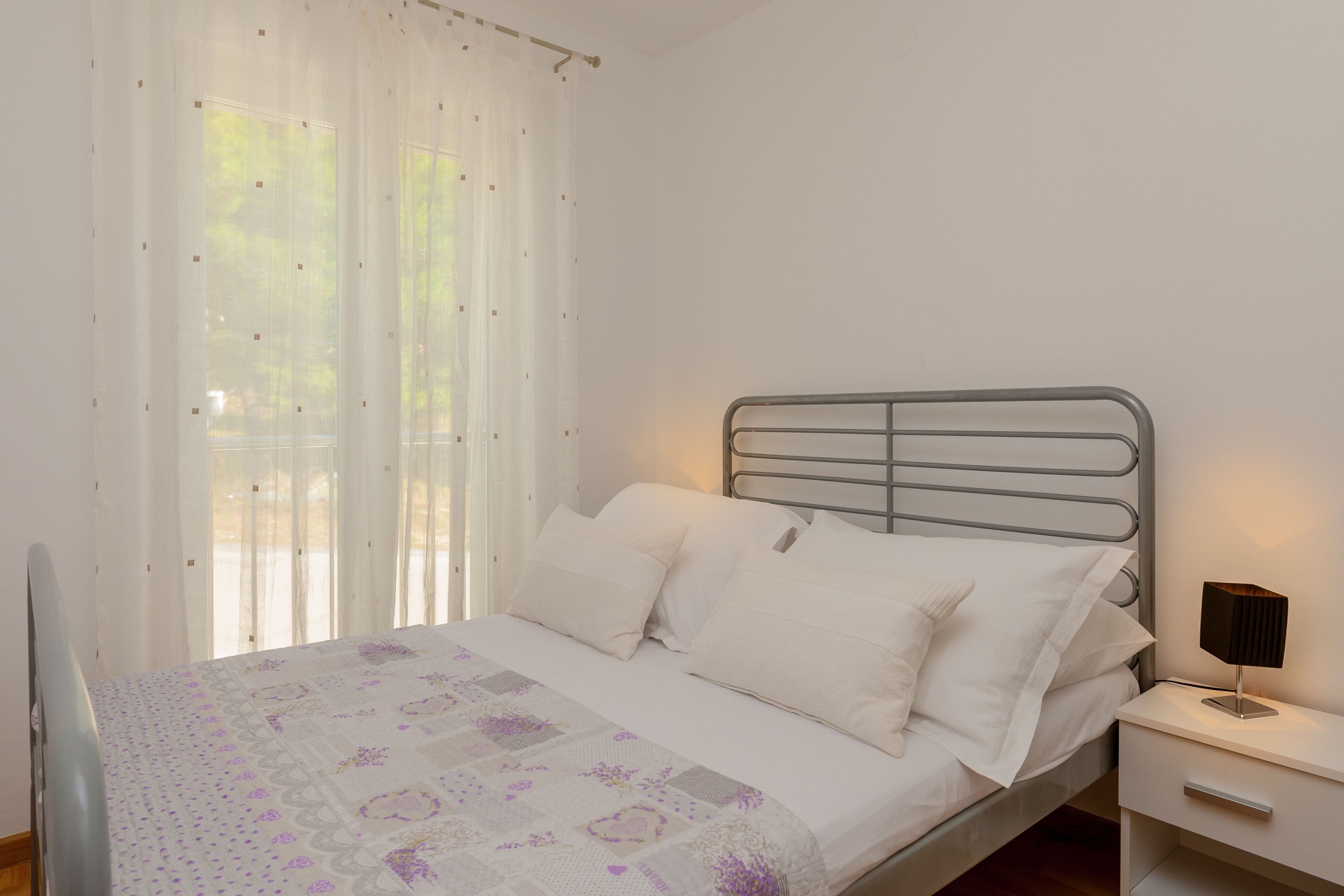 A neatly made bed with white linens and a floral-patterned blanket, a bedside table with a lamp, and sheer curtains allowing natural light.