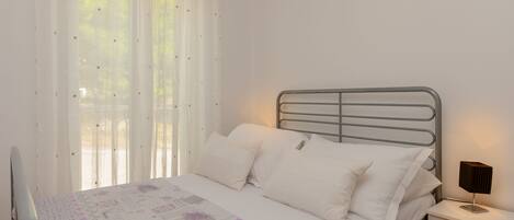 A neatly made bed with white linens and a floral-patterned blanket, a bedside table with a lamp, and sheer curtains allowing natural light.