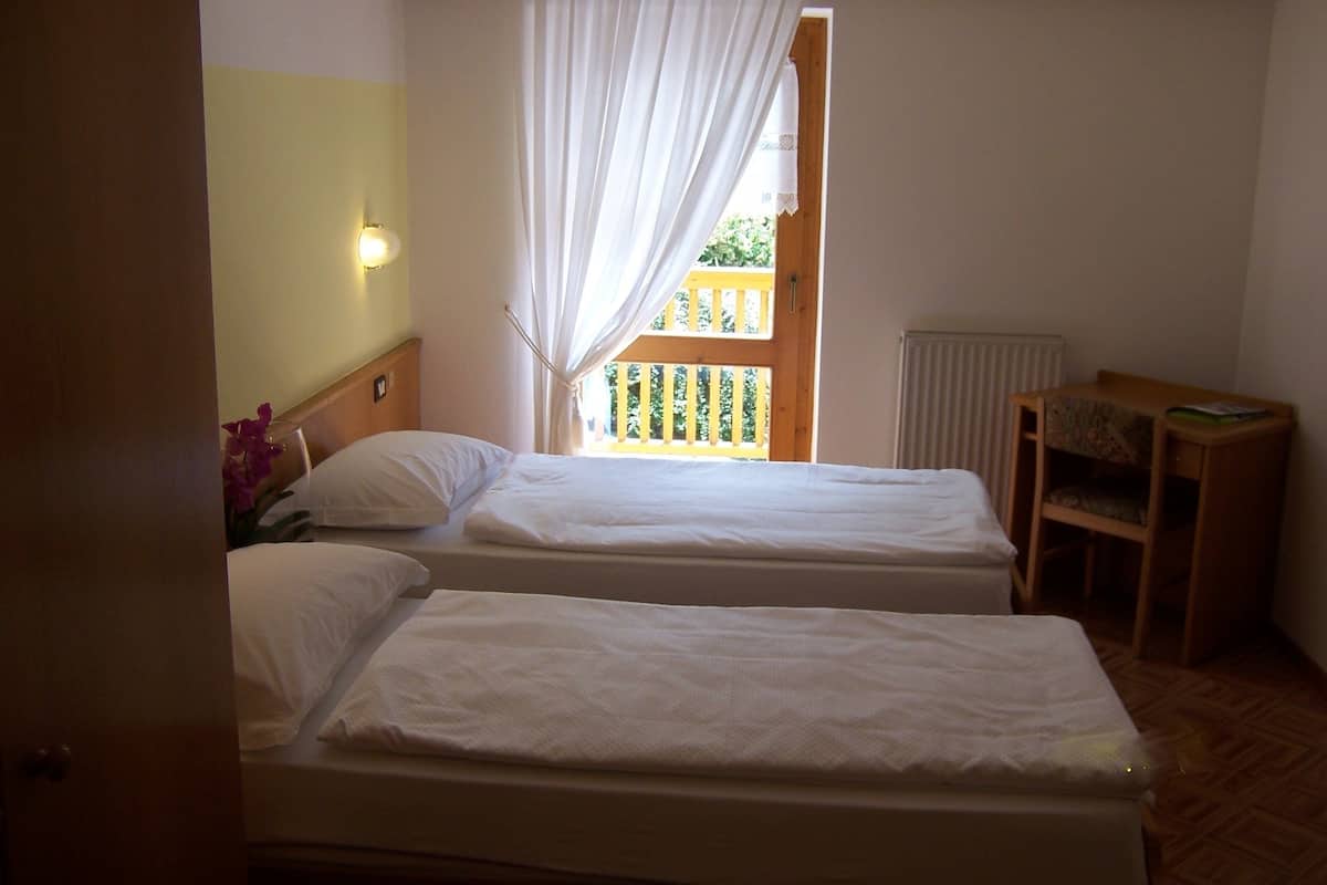 A room with two single beds, a wooden headboard, a small table with a chair, and a window with white curtains.