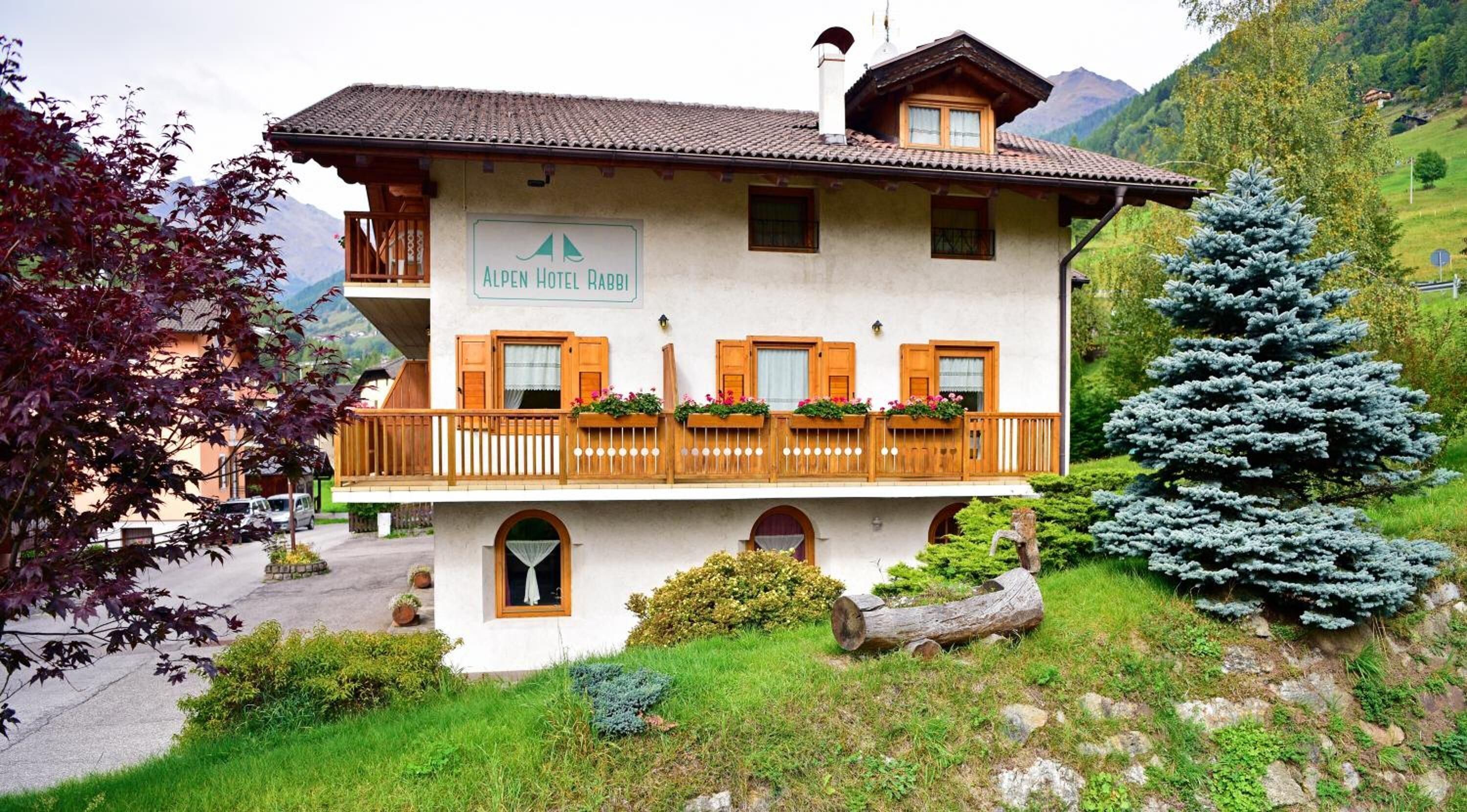 Un edificio de dos pisos con un letrero que dice "Alpen Hotel Rabbi", con un balcón con barandillas de madera y macetas con flores, rodeado de vegetación y montañas.
