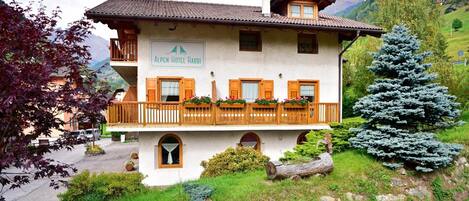 Un edificio de dos pisos con un letrero que dice "Alpen Hotel Rabbi", con un balcón con barandillas de madera y macetas con flores, rodeado de vegetación y montañas.