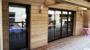 Ein Holzfassade mit einer Schiebetür, einer überdachten Terrassenfläche mit Stühlen und einem kleinen rosafarbenen Tisch.