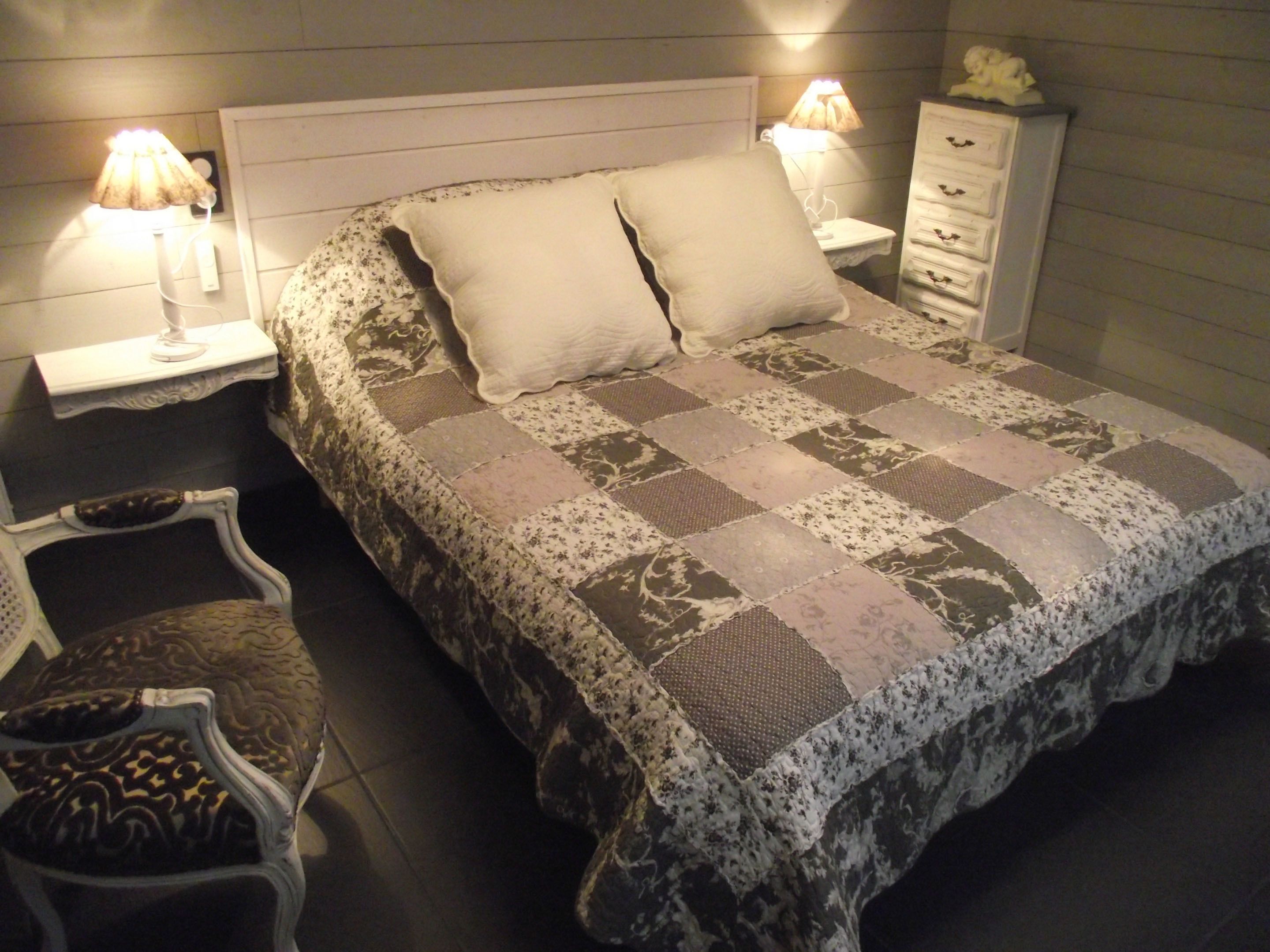 Un lit bien fait, recouvert d’une courtepointe, avec deux lampes de chevet, une table de chevet avec une lampe, une commode avec des tiroirs et une chaise agrémentée d’un coussin à motifs.