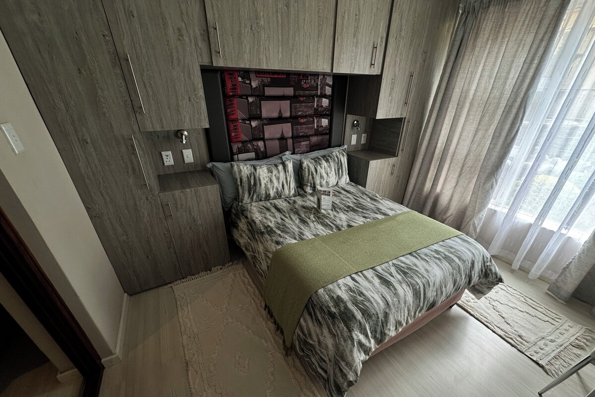 Une chambre à coucher comprenant un lit, une tête de lit ornée d’un motif représentant un paysage urbain, une table de chevet, une commode, une fenêtre avec des rideaux et un plancher en bois.