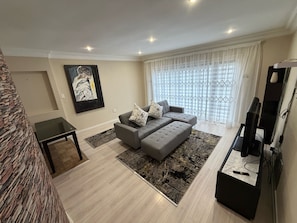 Un salon moderne avec un canapé d’angle gris, un téléviseur à écran plat et une grande fenêtre ornée de rideaux légers.
