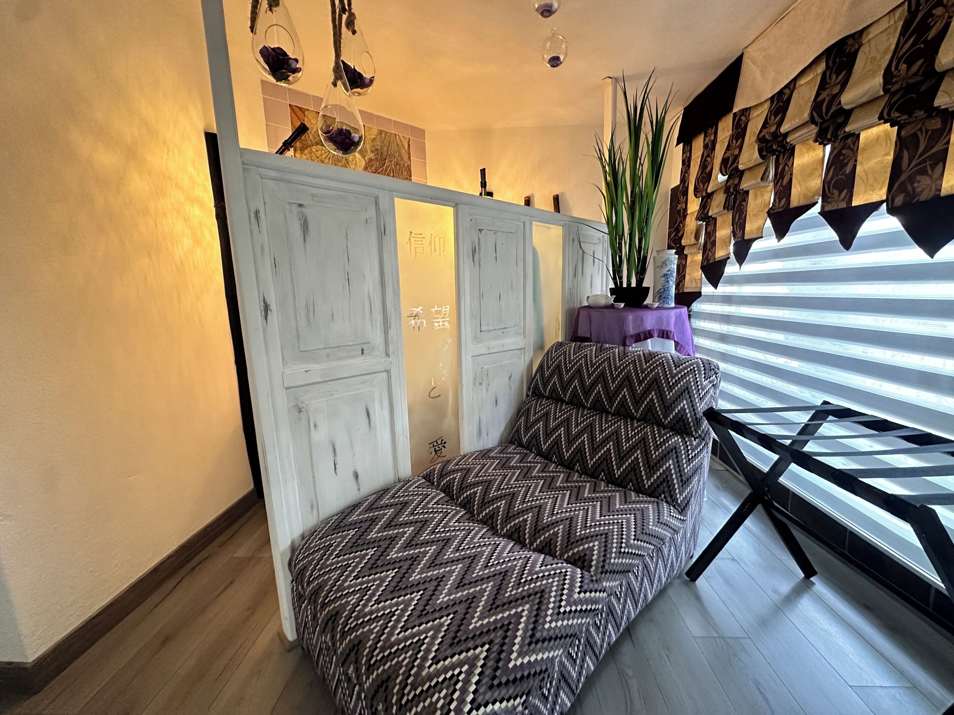 Een kamer met een gestreept bankstel, een witte garderobe, een potplant en een raam met zonwering.