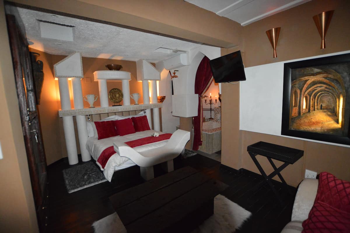 Habitación de hotel con cama, una mesita, televisor de pared y un cuadro que muestra un pasillo arquitectónico.