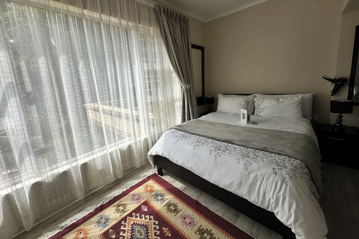 Une chambre d’hôtel avec un lit, une table de chevet, une lampe, une fenêtre avec des rideaux légers et un tapis à motifs.