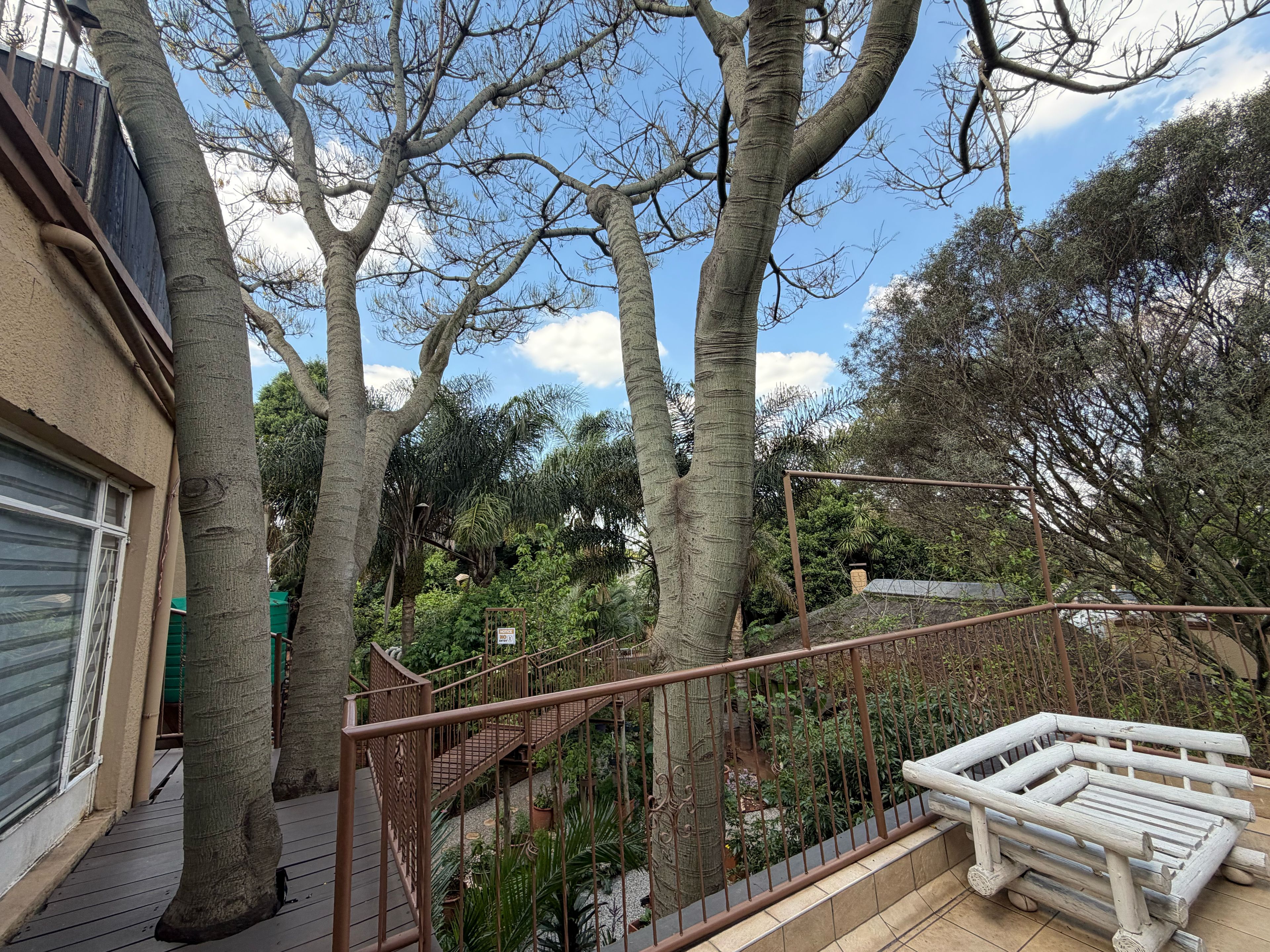 Un balcón con barandilla de madera, vista a un jardín con diversas plantas y un edificio con puerta de garaje.