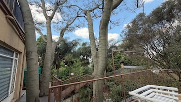 Un balcón con barandilla de madera, vista a un jardín con diversas plantas y un edificio con puerta de garaje.