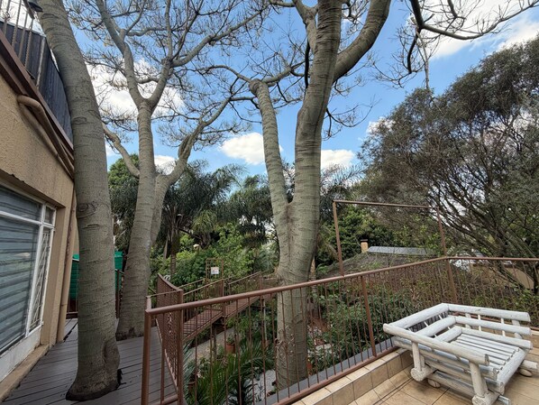 Un balcon avec une balustrade en bois, une vue sur un jardin planté de différentes espèces, et un bâtiment doté d’une porte de garage.