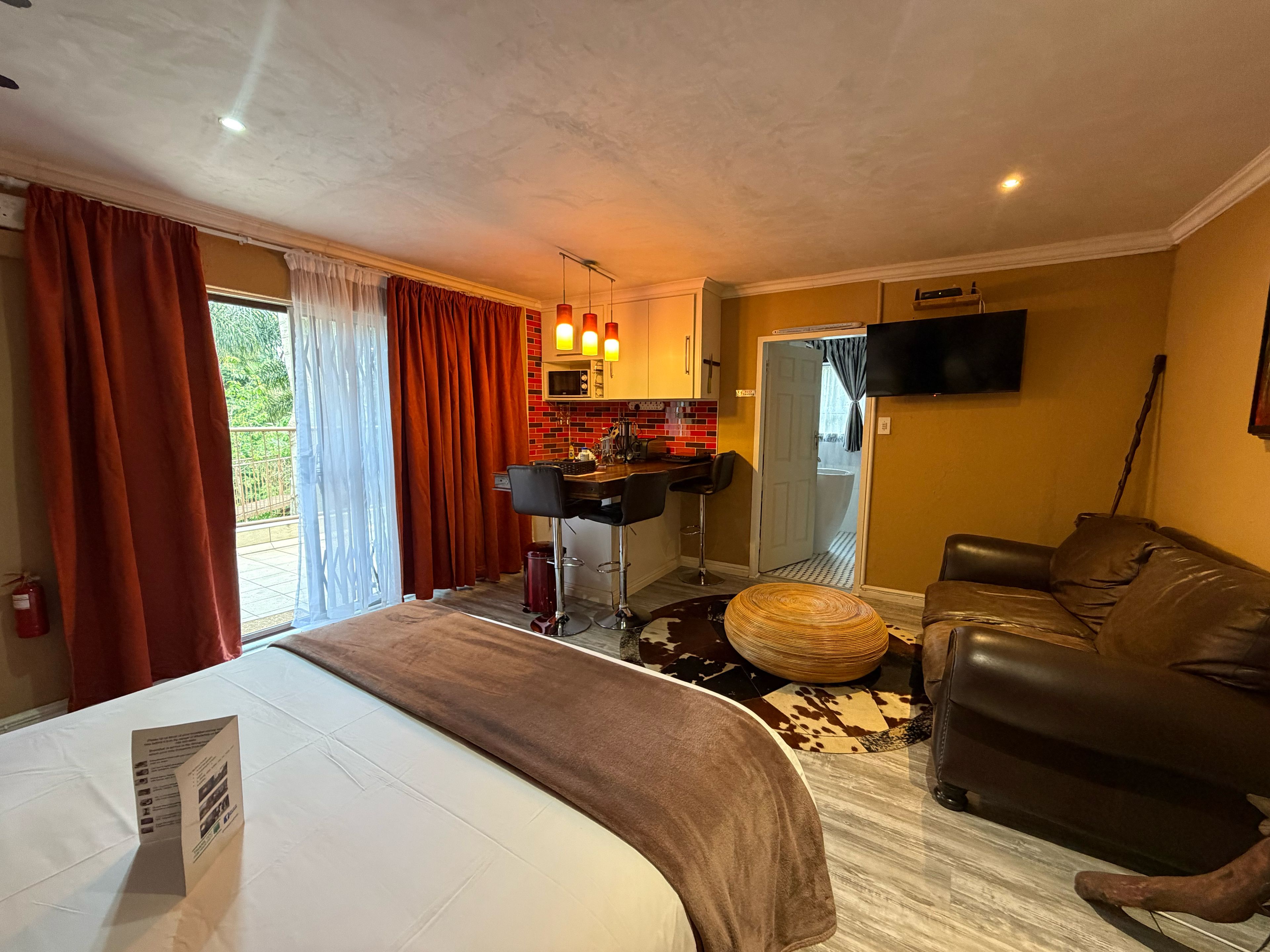 A hotel room with a bed, a sofa, a small kitchen area, and a TV mounted on the wall.