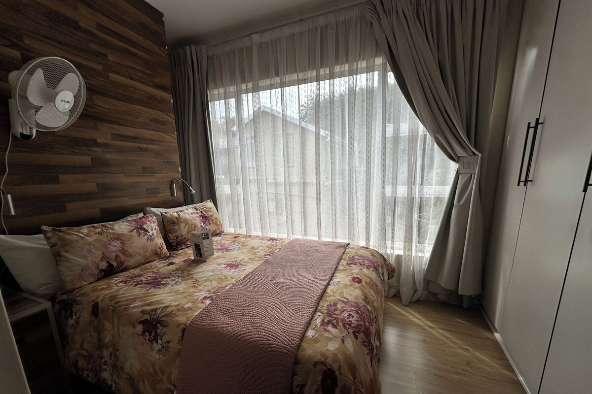 Une chambre avec une tête de lit en bois, un lit recouvert d’une literie à motifs floraux, un ventilateur sur pied et une fenêtre ornée de rideaux légers.