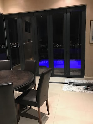 A dining area with a table and chairs, a pool with blue lighting, and a view of the city at night.