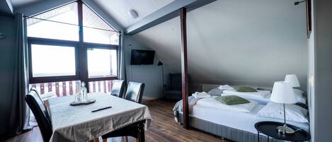 A room with a dining table, chairs, a bed with pillows, a TV, and a window.
