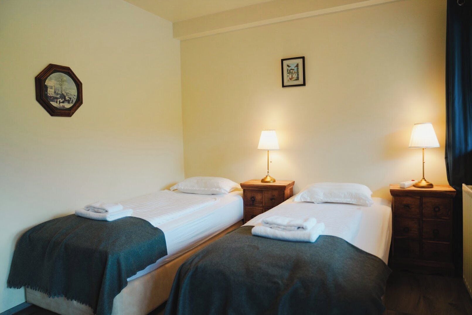 Habitación de hotel con dos camas individuales, cada una con mesita de noche y lámpara. Encima de las camas hay un cuadro en la pared.