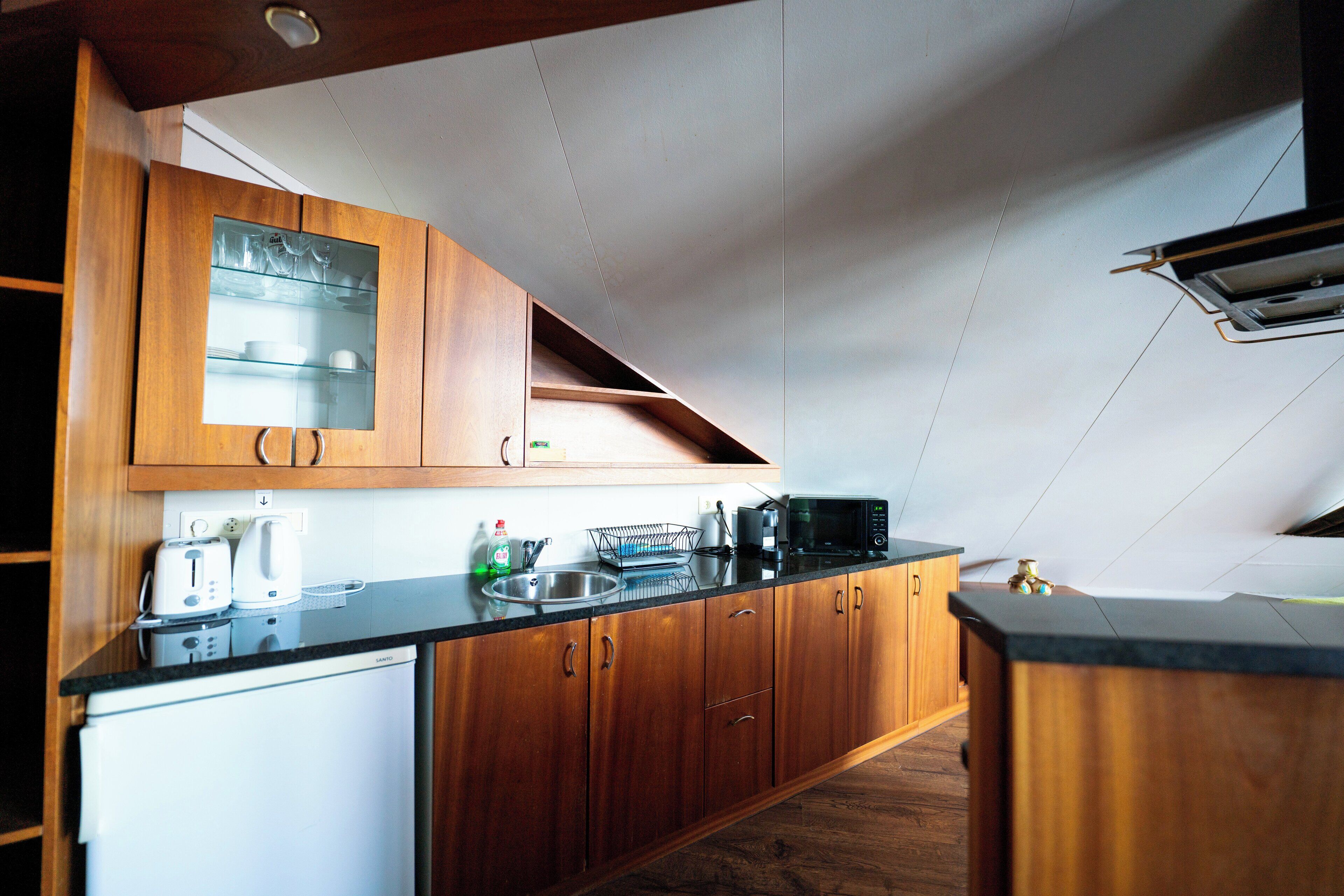 Una cocina con armarios de madera, encimera negra, microondas y un refrigerador pequeño.