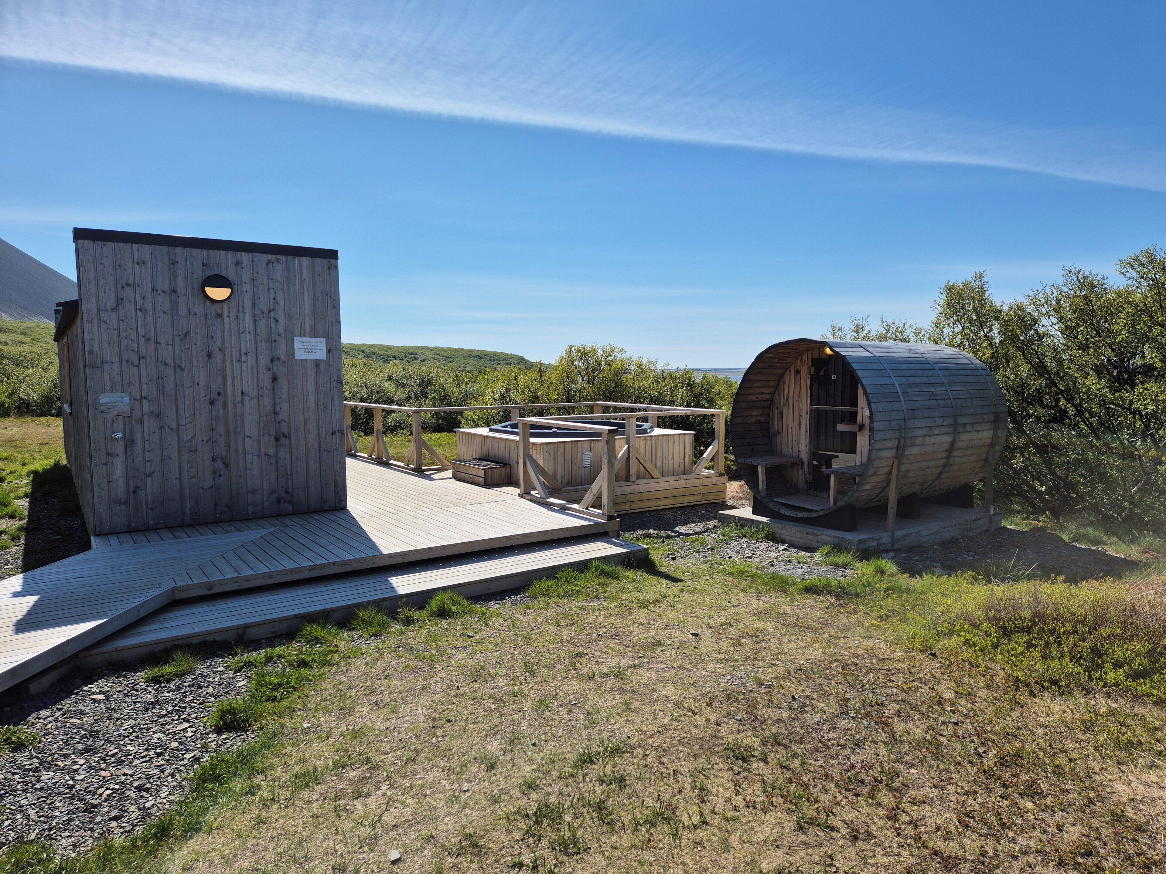 A wooden cabin with a hot tub and a barrel-shaped structure, set in a grassy area with a clear sky.