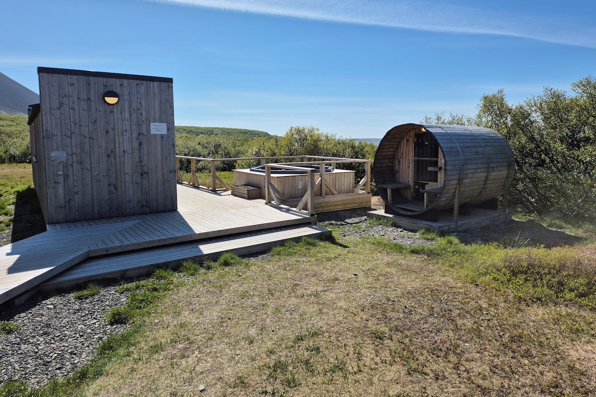 A wooden cabin with a hot tub and a barrel-shaped structure, set in a grassy area with a clear sky.