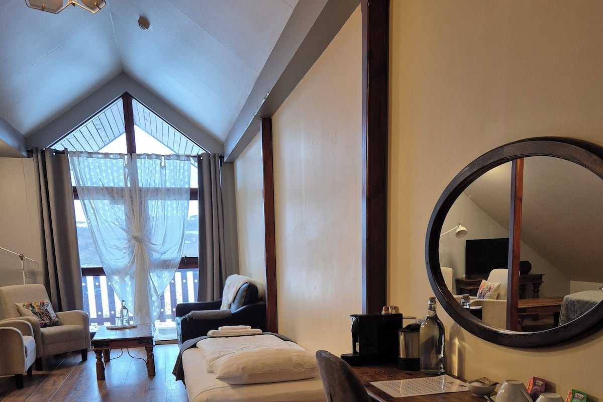 A hotel room with a wooden floor, a bed, a desk with a mirror, a chair, and a window with curtains.