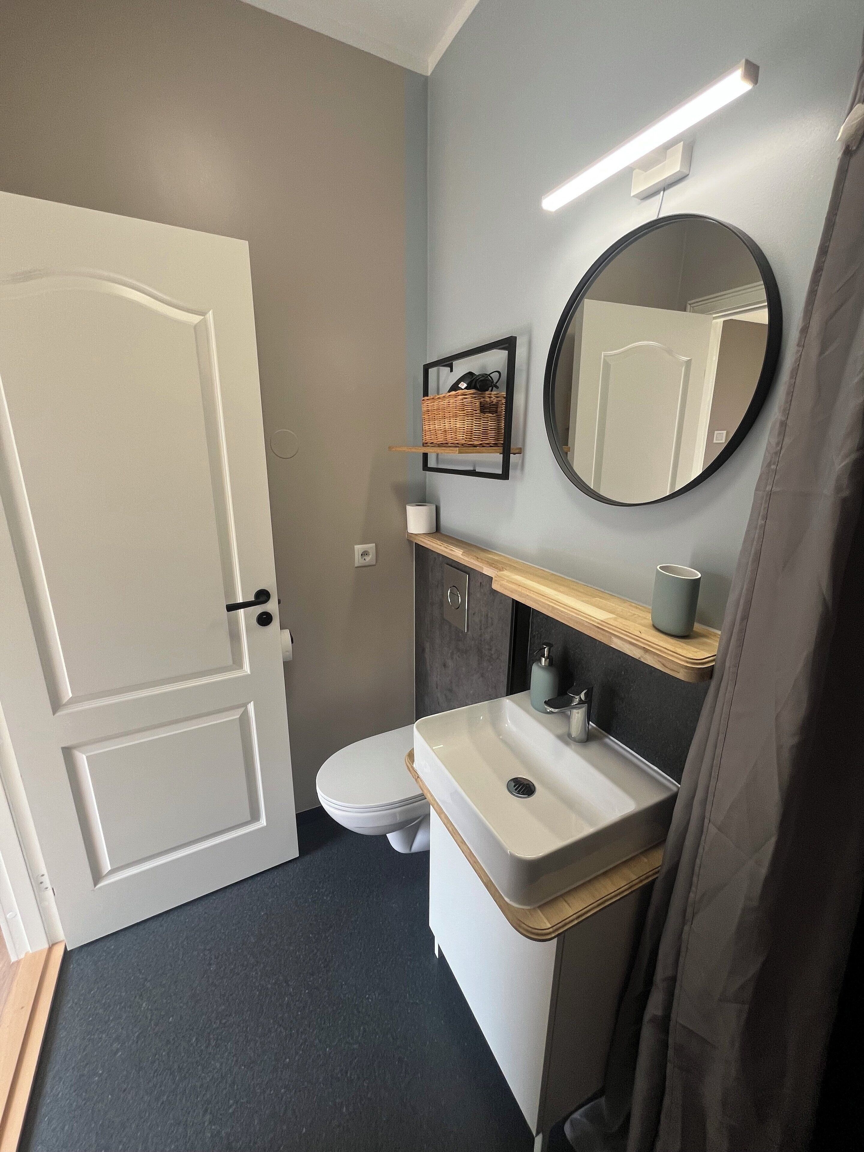 Een badkamer met een witte deur, een toilet, een wastafel met spiegel en een plank met een mand en een beker.