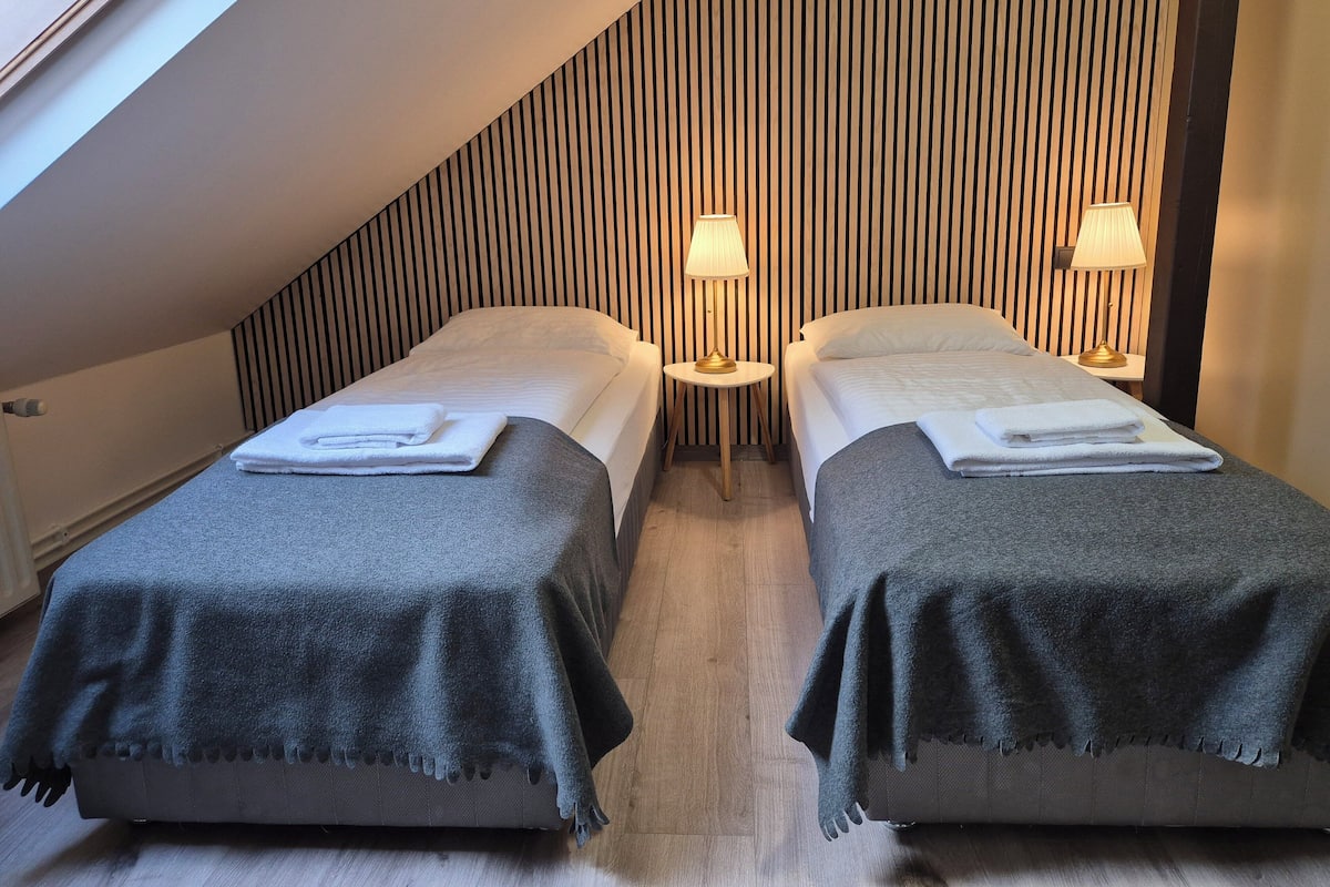 A room with two beds, each with a grey blanket and white towels, a striped wall, and two bedside lamps.