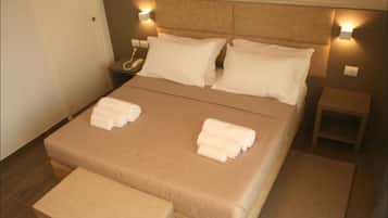 A neatly made bed with white linens and two towels folded on the bed. There are two bedside tables, one with a phone and the other with a lamp. The room has a wooden floor and a small bench at the foot of the bed.