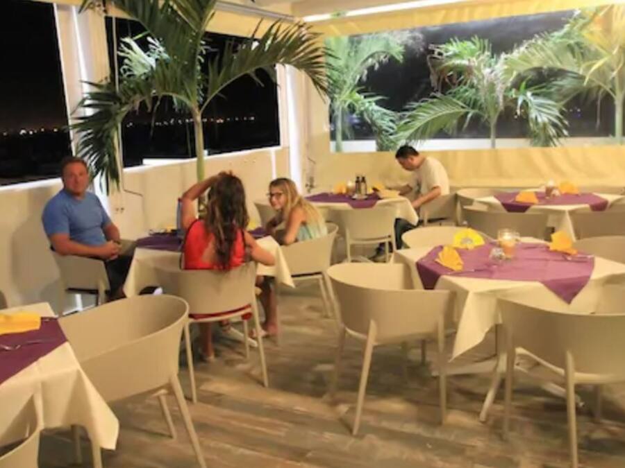 Un área de comedor al aire libre con mesas y sillas redondas, palmeras y vistas de la ciudad de noche.