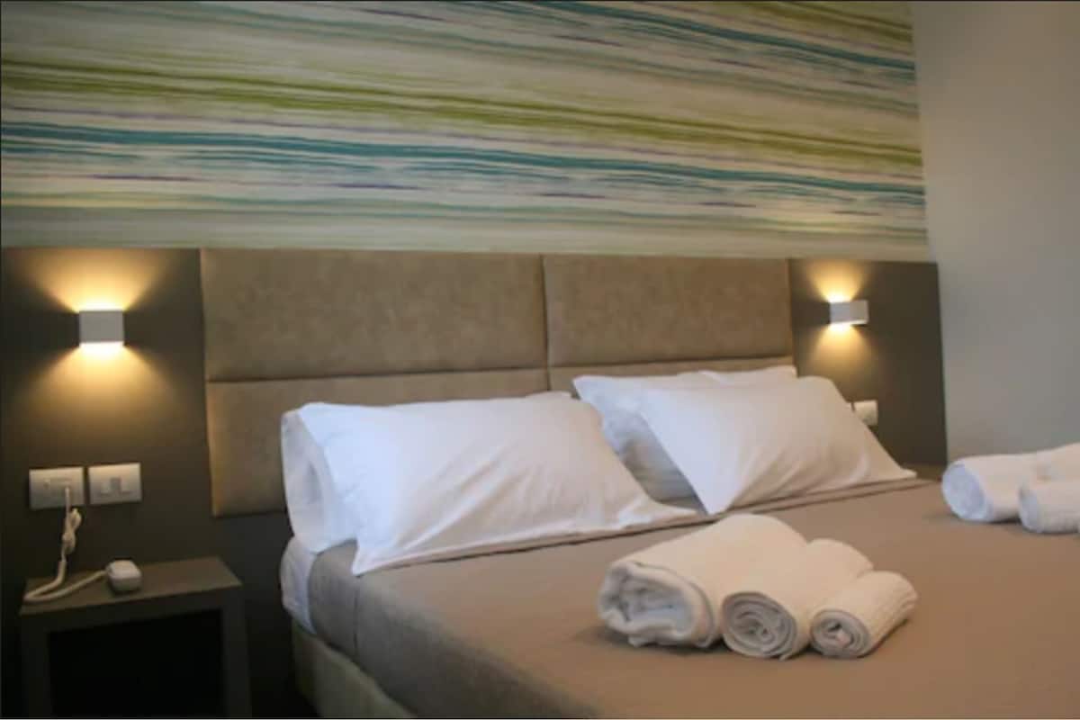 A hotel room with a large bed, two pillows, a headboard with wall-mounted lights, a bedside table with a phone, and rolled towels on the bed.
