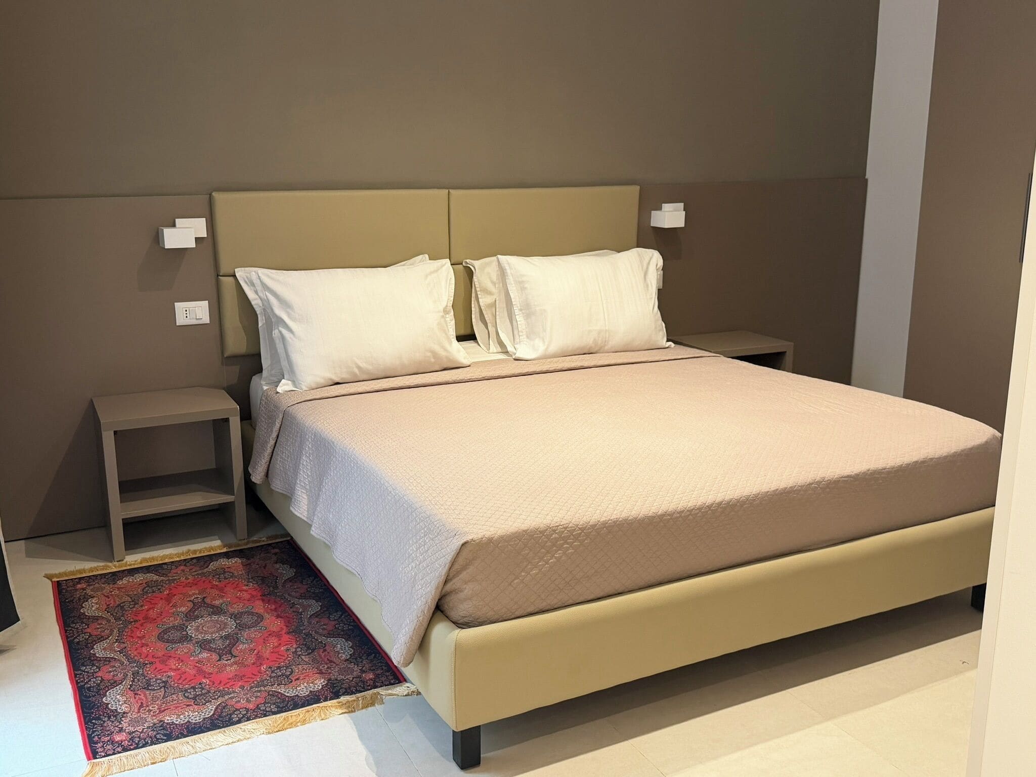 A bedroom with a bed, nightstands, a bedside table, and a patterned rug.