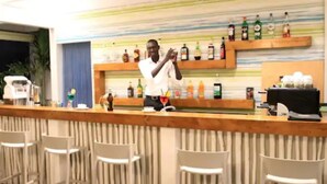 Una zona de bar con mostrador de madera, sillas blancas y una pared con estantes llenos de botellas.
