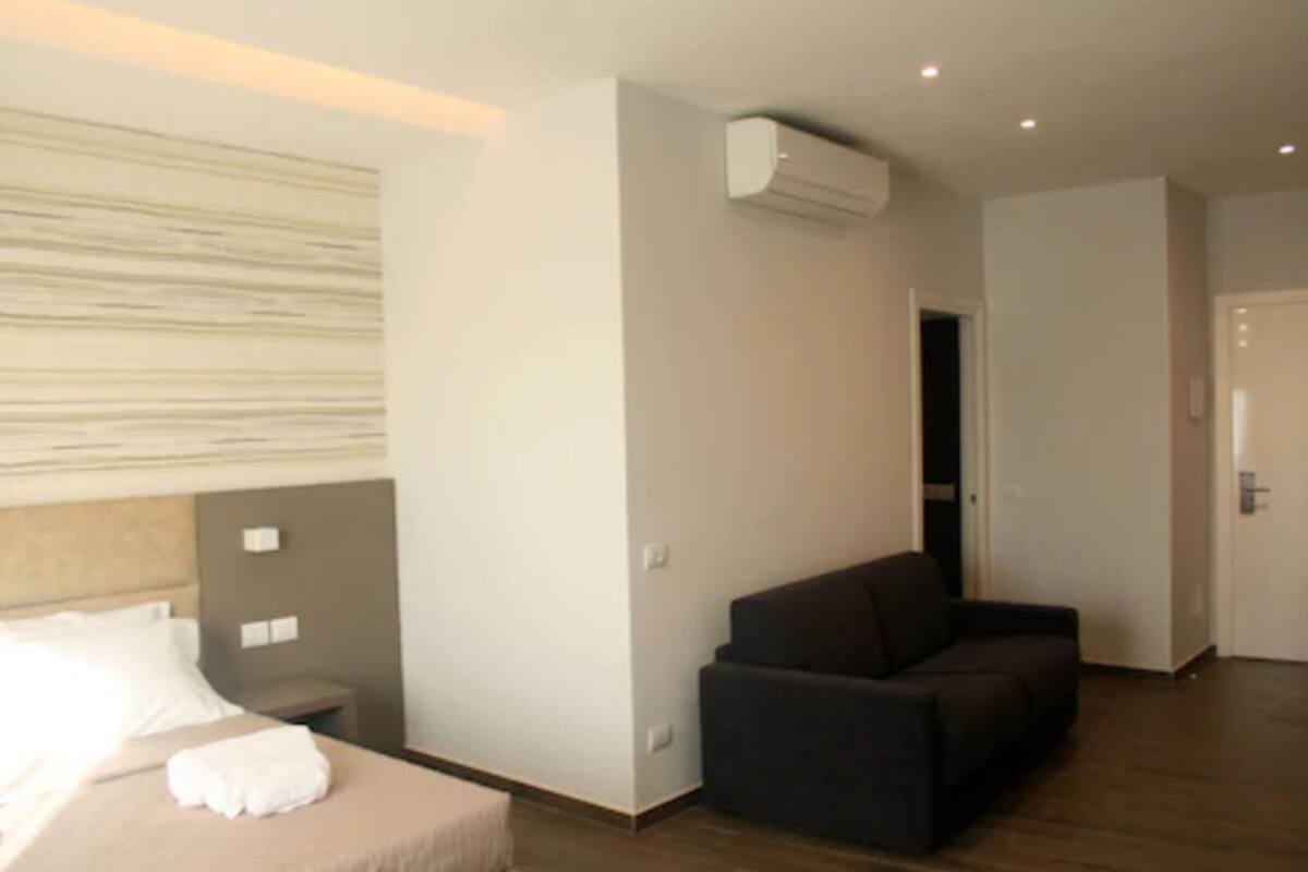 A modern hotel room with a bed, a dark sofa, and an air conditioning unit.