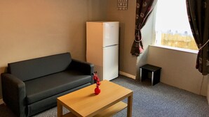 A small room with a dark sofa, a wooden table with a red vase, a white refrigerator, and a window with floral curtains.