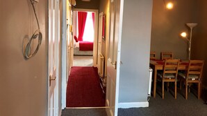 A hallway leading to a bedroom with a red carpet, a dining area with wooden chairs, and a lamp.