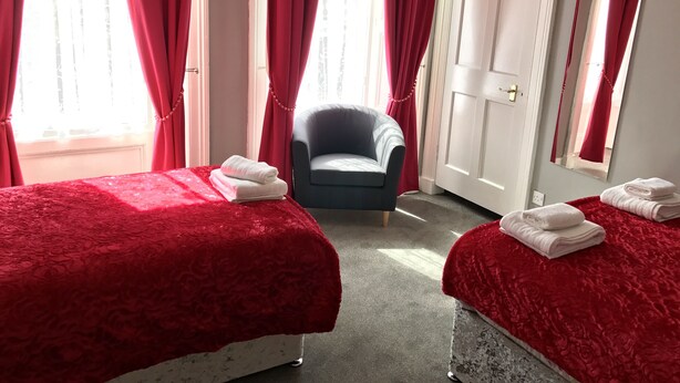 A hotel room with two beds, each with a red bedspread and white towels, a grey armchair, a white door, and a mirror on the wall.