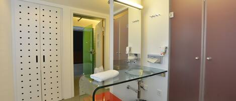 Un baño moderno con un lavamanos de vidrio, una toalla blanca, un cubo de basura y un armario blanco con diseño perforado.