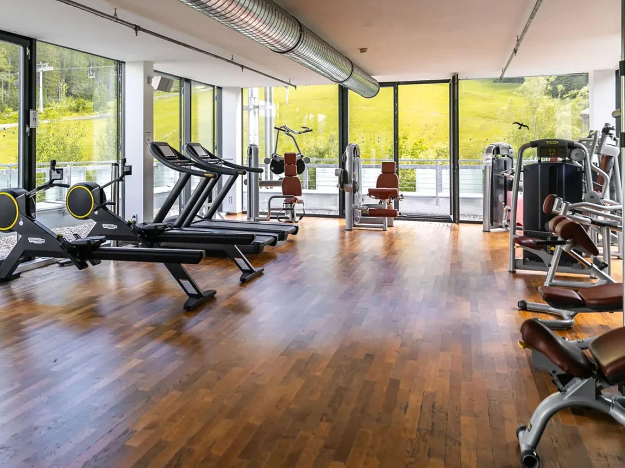 Un gimnasio con máquinas de cardio, estaciones de pesas y un amplio ventanal con vistas a zonas verdes.